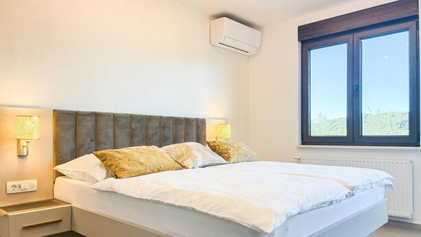 A bedroom with a bed, bedside lamps, an air conditioning unit, a window, and a radiator. - Cozy apartment in Palit with WiFi (Palit)
