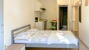Un dormitorio con cama, mesita de noche, escritorio pequeño, silla, baño con ducha y zona de cocina.