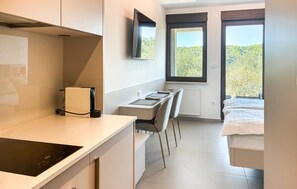Ein modernes Hotelzimmer mit Küchenzeile, einem Bett, einem Schreibtisch und einem großen Fenster mit Blick auf Grünflächen.