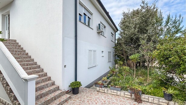 Un bâtiment à deux étages, à l’extérieur blanc, avec un escalier menant à l’entrée et un jardin agrémenté de diverses plantes.
