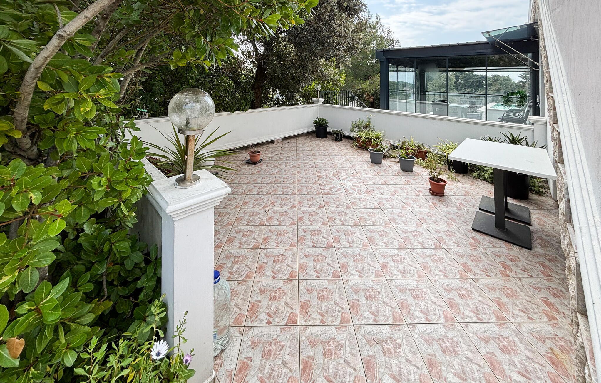 Terraza en la azotea con plantas en macetas, barandilla de cristal y una mesa blanca.