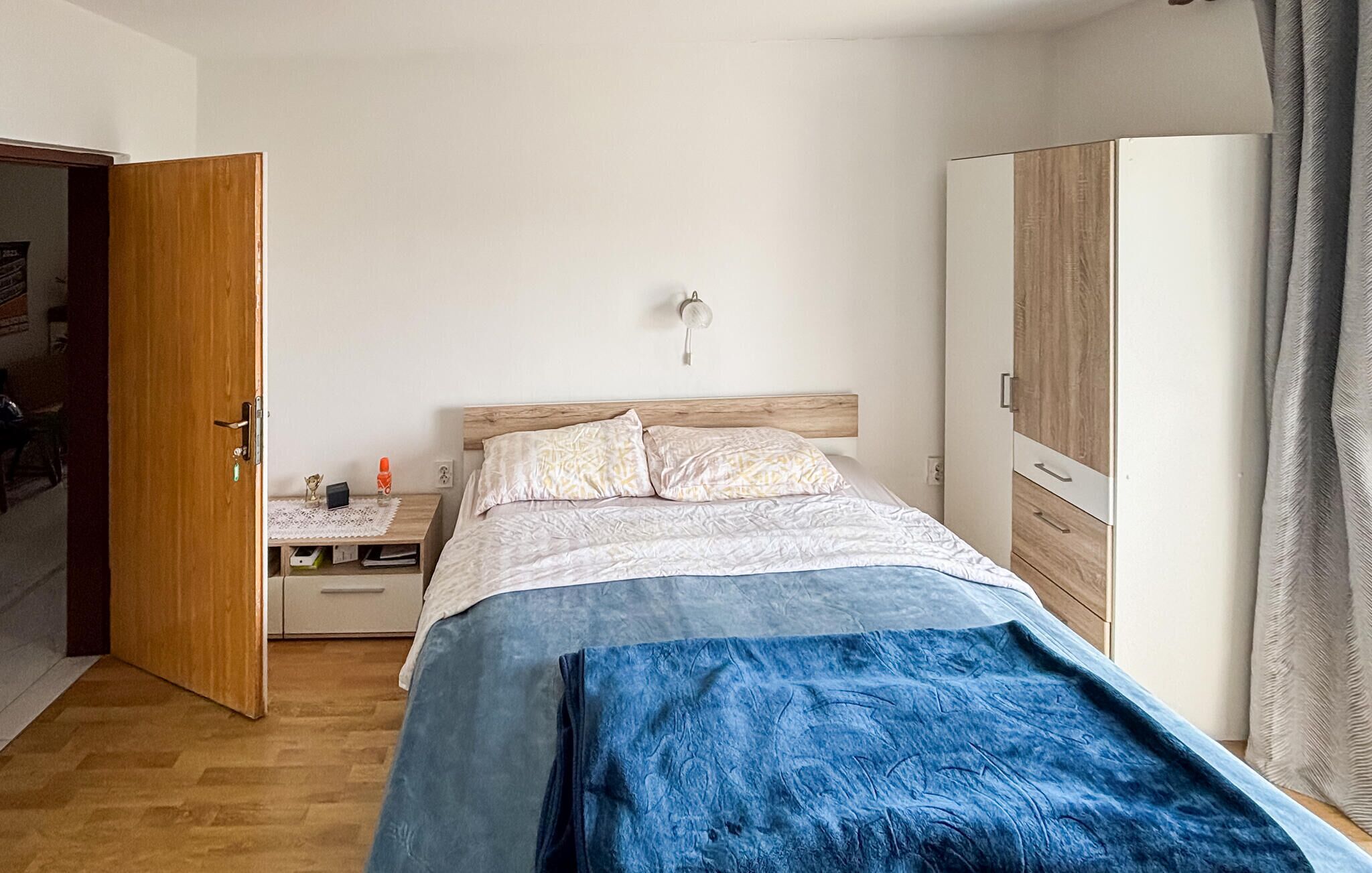 Un dormitorio con cama, mesita de noche, lámpara, armario y una puerta abierta que da a otra habitación.