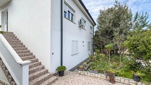 A two-story building with a white exterior, a set of stairs leading to the entrance, and a garden area with various plants. - Nice apartment in Rab with WiFi (Rab)