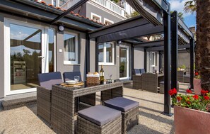 Un espace extérieur aménagé en terrasse, avec des meubles en osier, une table sur laquelle se trouvent une bouteille et des verres, et une vue sur un bâtiment doté de grandes fenêtres.