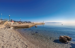 Ein Kiesstrand mit einem Steg, klarem, blauem Wasser und fernen Bergen.
