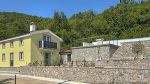 Un edifici groc amb un balcó, una paret de pedra i un vessant boscoso al fons.