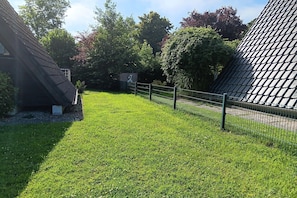 A well-maintained grassy area with a metal fence, surrounded by trees and a building with a sloped roof. - gemütliche Unterkunft in Burhave (Fedderwardersiel)