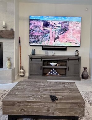 Un salon doté d’un téléviseur à écran plat fixé au-dessus d’un meuble de rangement gris, d’une table basse en bois placée au premier plan et d’une cheminée agrémentée d’objets décoratifs.