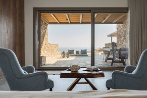 Habitación con un amplio ventanal corredero que da a un patio exterior con pared de piedra y vista al mar. Hay dos sillas y una mesita con un cuenco y un libro encima.