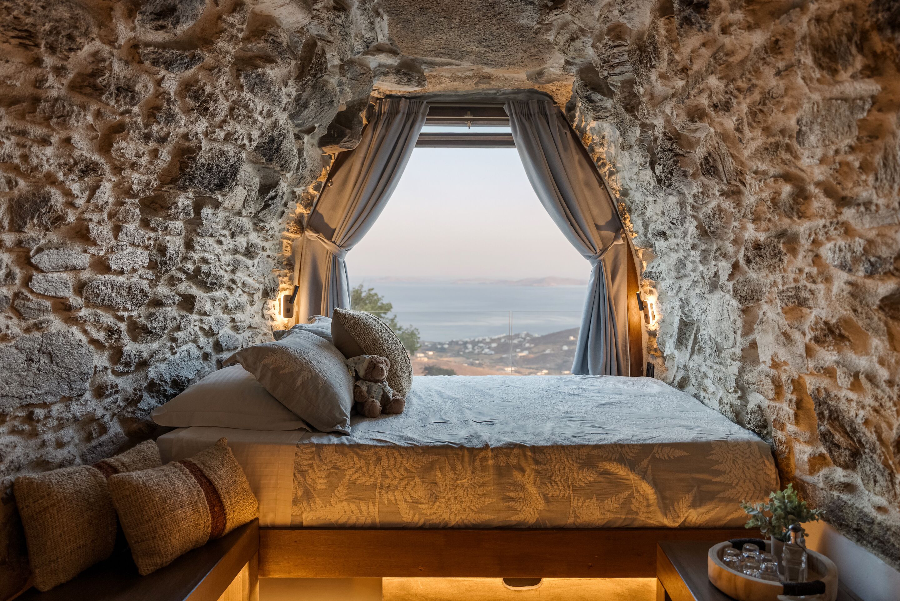 A bedroom with a stone wall, a bed with pillows, a teddy bear, a window with curtains, and a view of the sea.