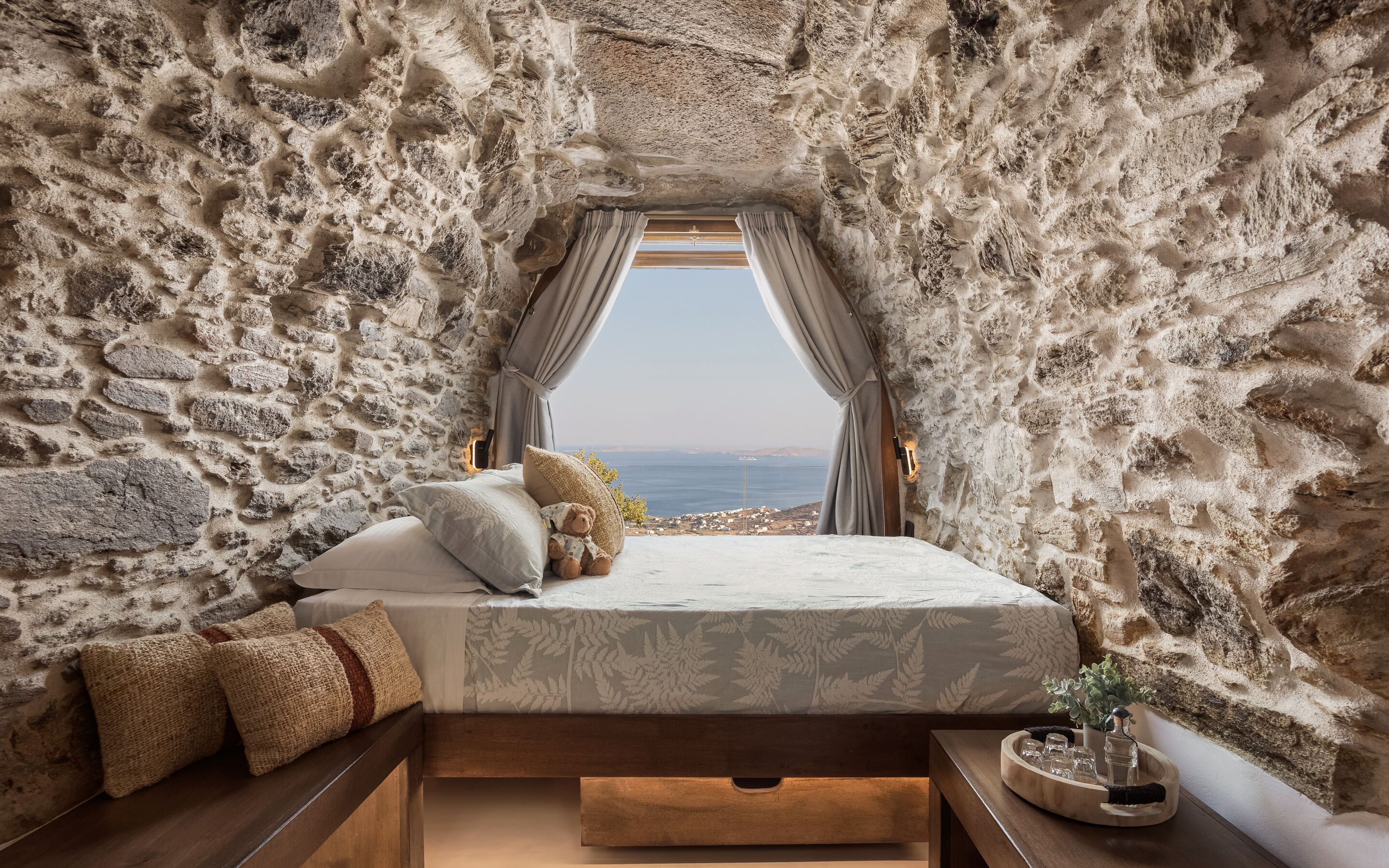 Un dormitorio con pared de piedra, una cama con colcha blanca y vistas al mar.