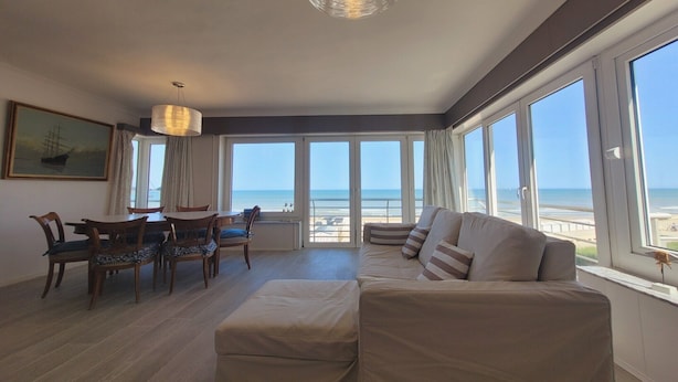 Ein geräumiges Wohnzimmer mit großem Fenster zum Strand, einem Essbereich mit Holzmöbeln und einer modernen Kronleuchter.