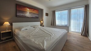 Ein modernes Hotelzimmer mit einem großen Bett, zwei Nachttischlampen, einem Bild über dem Bett, einem Fenster mit Vorhängen und Blick ins Freie.