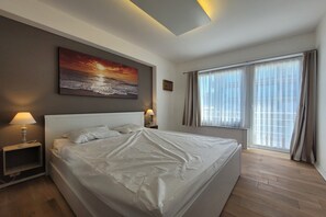 A modern hotel room with a large bed, two bedside lamps, a painting above the bed, a window with curtains, and a view of the outside. - Apartment with Panoramic Sea Views (MIDDELKERKE)