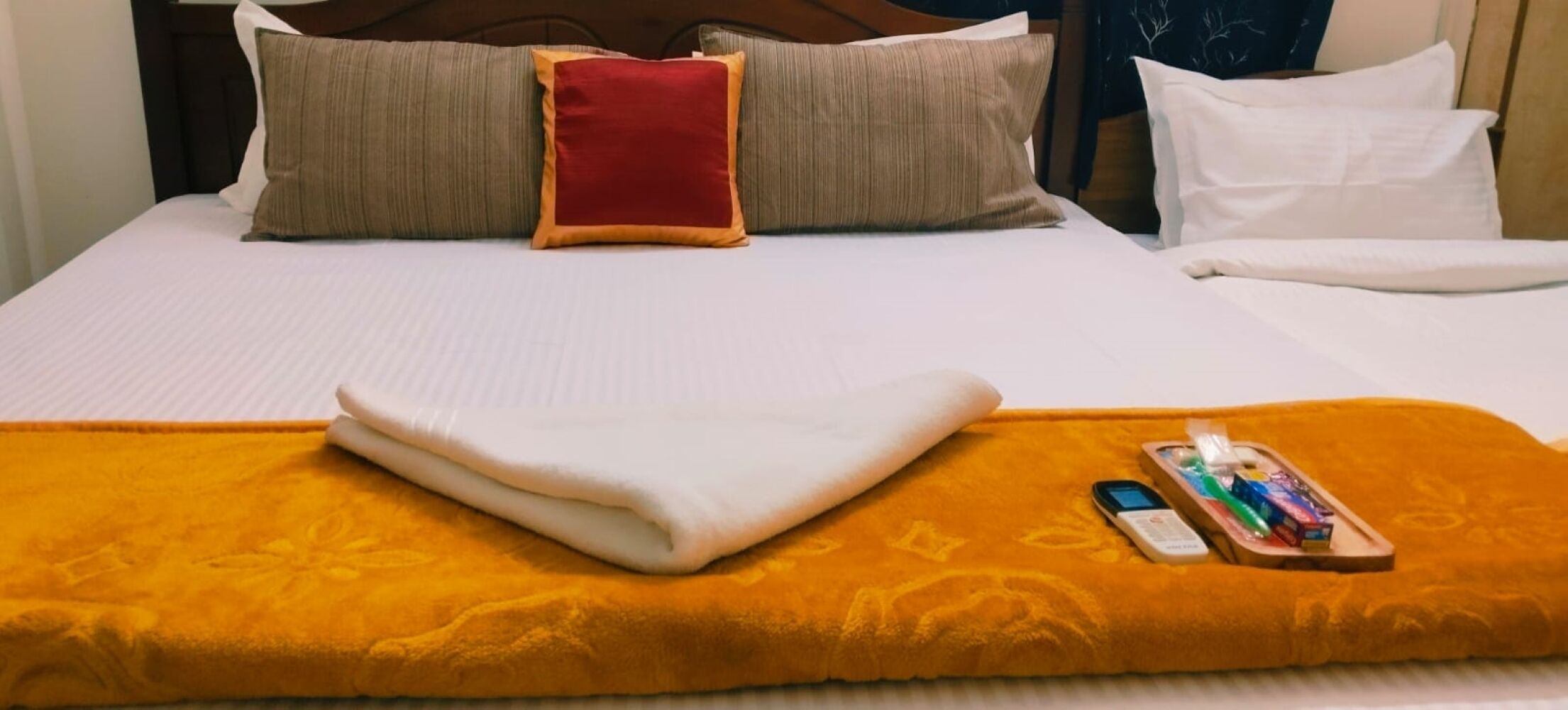 Una cama con una manta blanca y naranja, un cojín rojo y un teléfono inteligente con artículos de aseo sobre una bandeja.