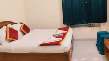 Una cama bien hecha con una colcha roja y blanca, almohadas y una toalla doblada. Junto a la cama hay una mesita de noche de madera con una planta pequeña y una tetera.