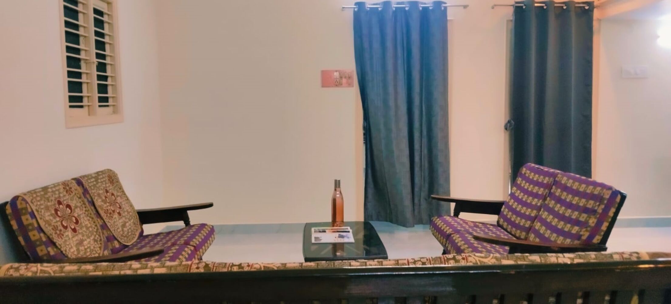 A living room with two patterned sofas, a small table with a bottle, and a window with blinds.