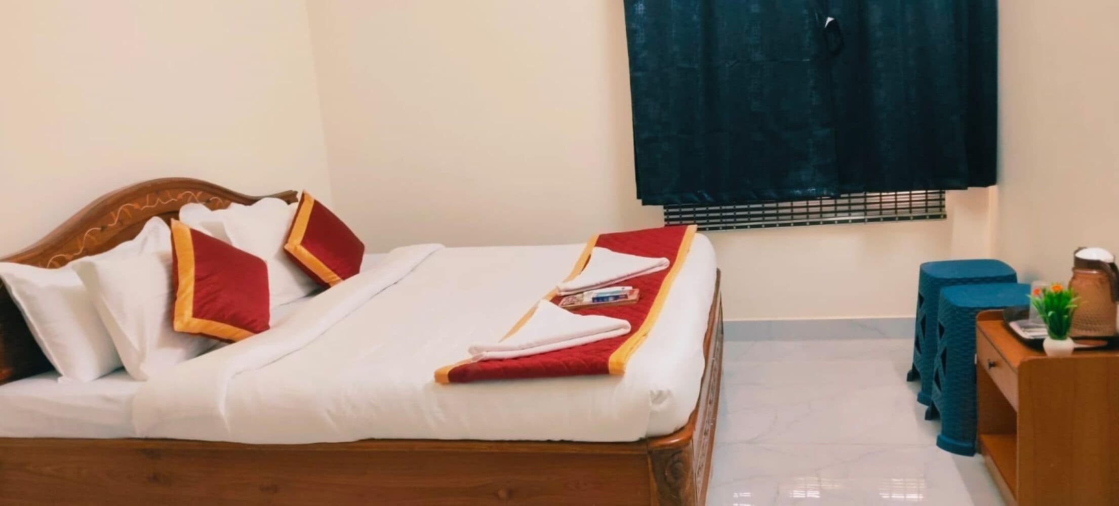A neatly made bed with white and red bedding, a wooden headboard, and a small bedside table with a kettle and a plant.