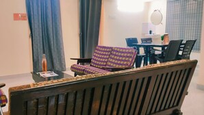 A room with a wooden sofa, two patterned armchairs, a small table with a bottle, and a dining area with chairs and a table. - Sarvik Thillai Residency  (Chidambaram)