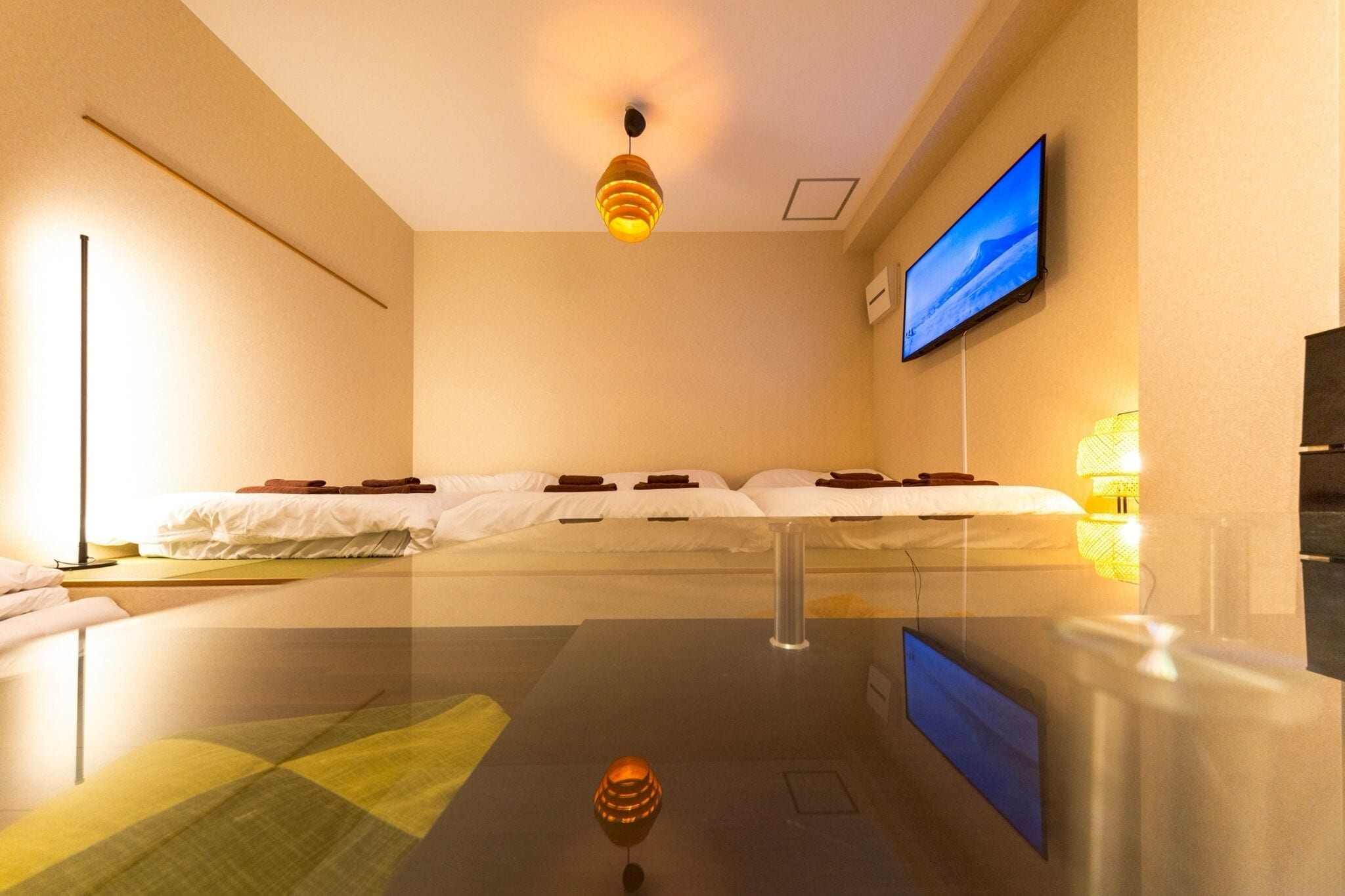 A hotel room with a bed, a flat-screen TV mounted on the wall, a ceiling light, and a glass table.