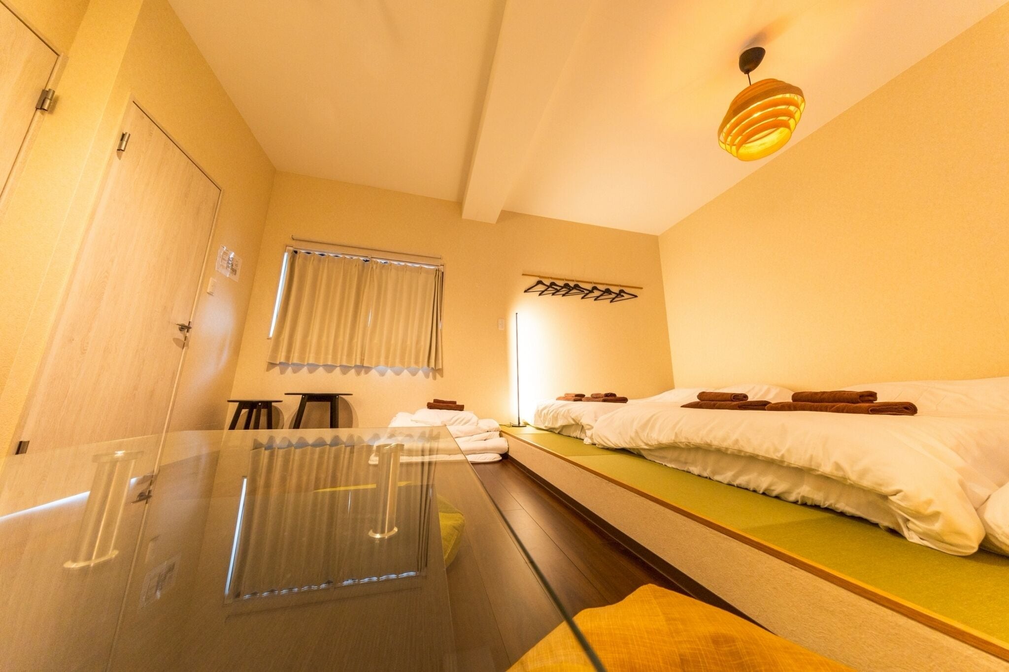 A hotel room with a glass table, a bed with white linens, a window with curtains, and a wall-mounted light fixture.