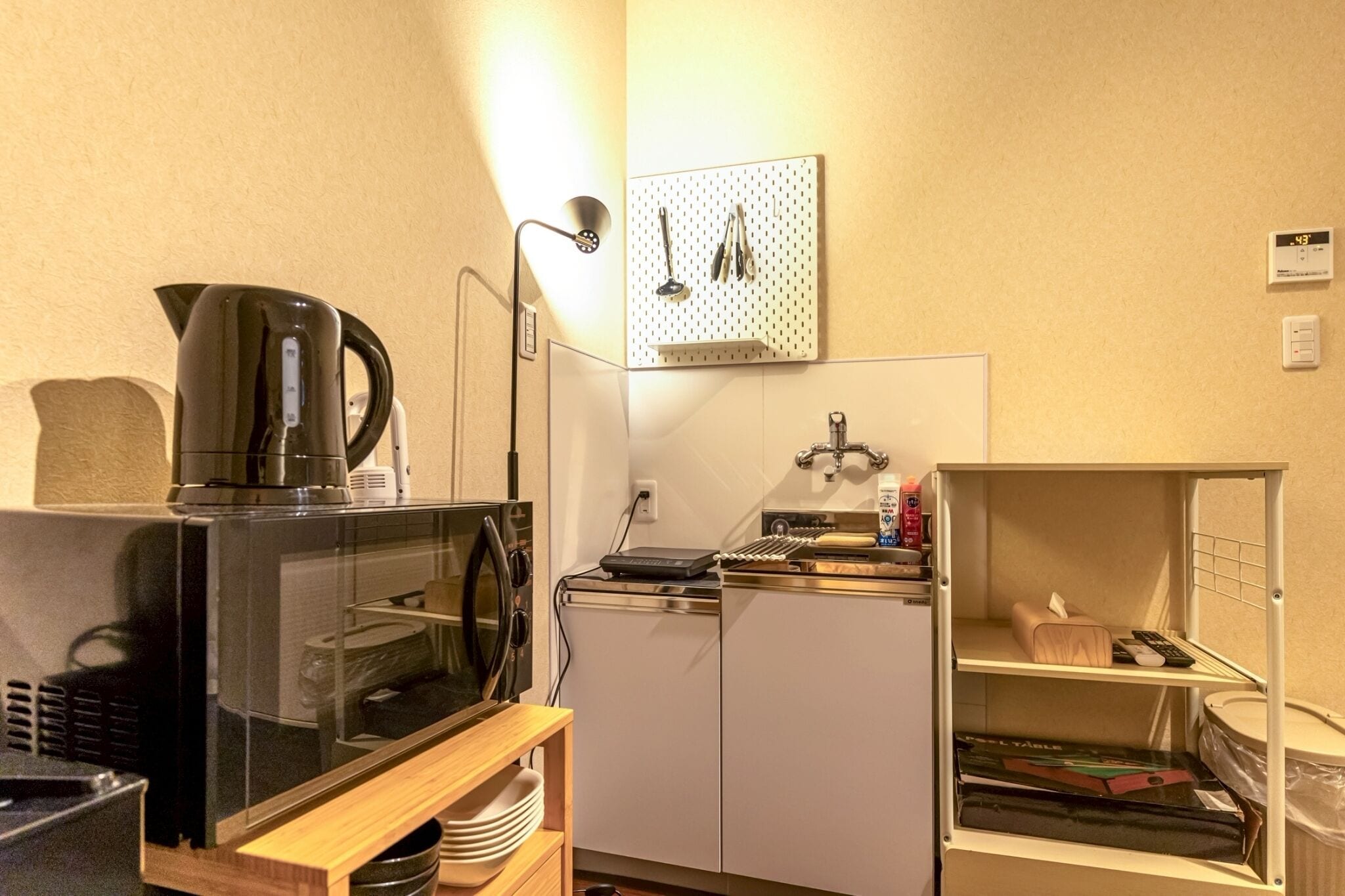 A kitchenette with a microwave, kettle, and coffee maker, a pegboard with kitchen utensils, a small refrigerator, and a shelf with a remote control and books.