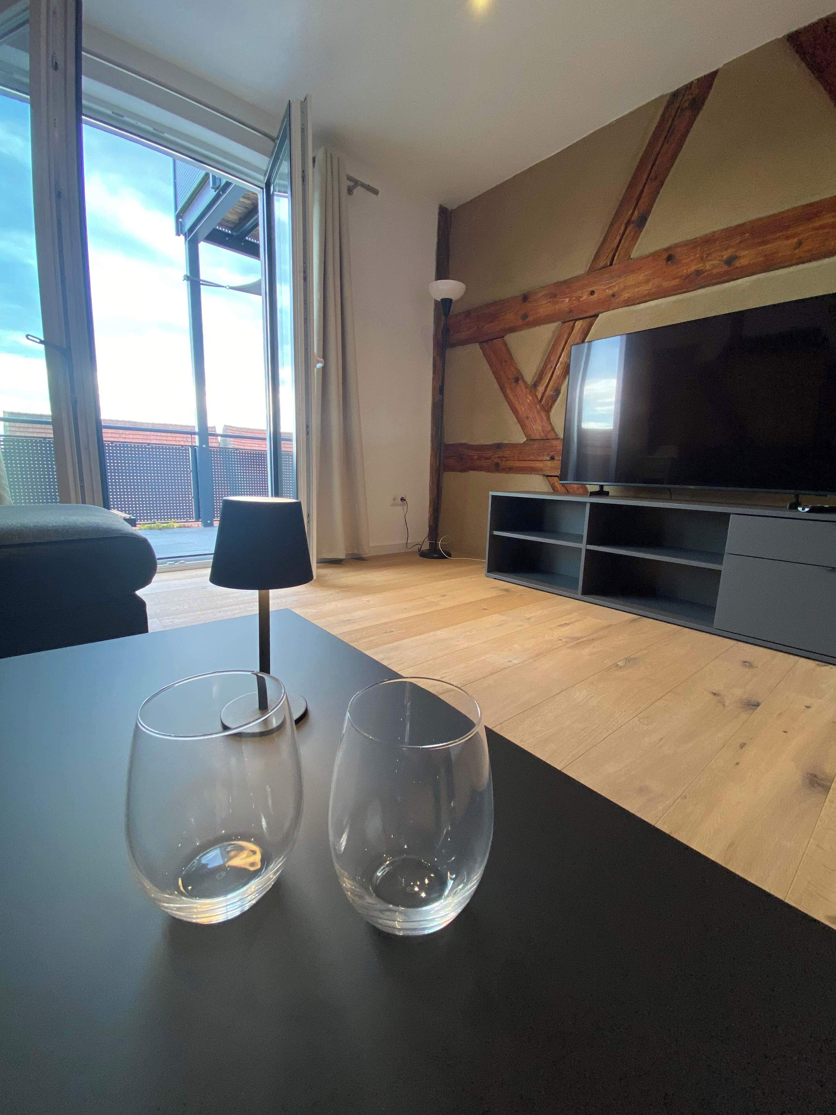 En moderne stue med stort fjernsyn, træbjælker, en balkon og to tomme glas på et bord.