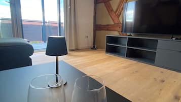 En moderne stue med stort fjernsyn, træbjælker, en balkon og to tomme glas på et bord.