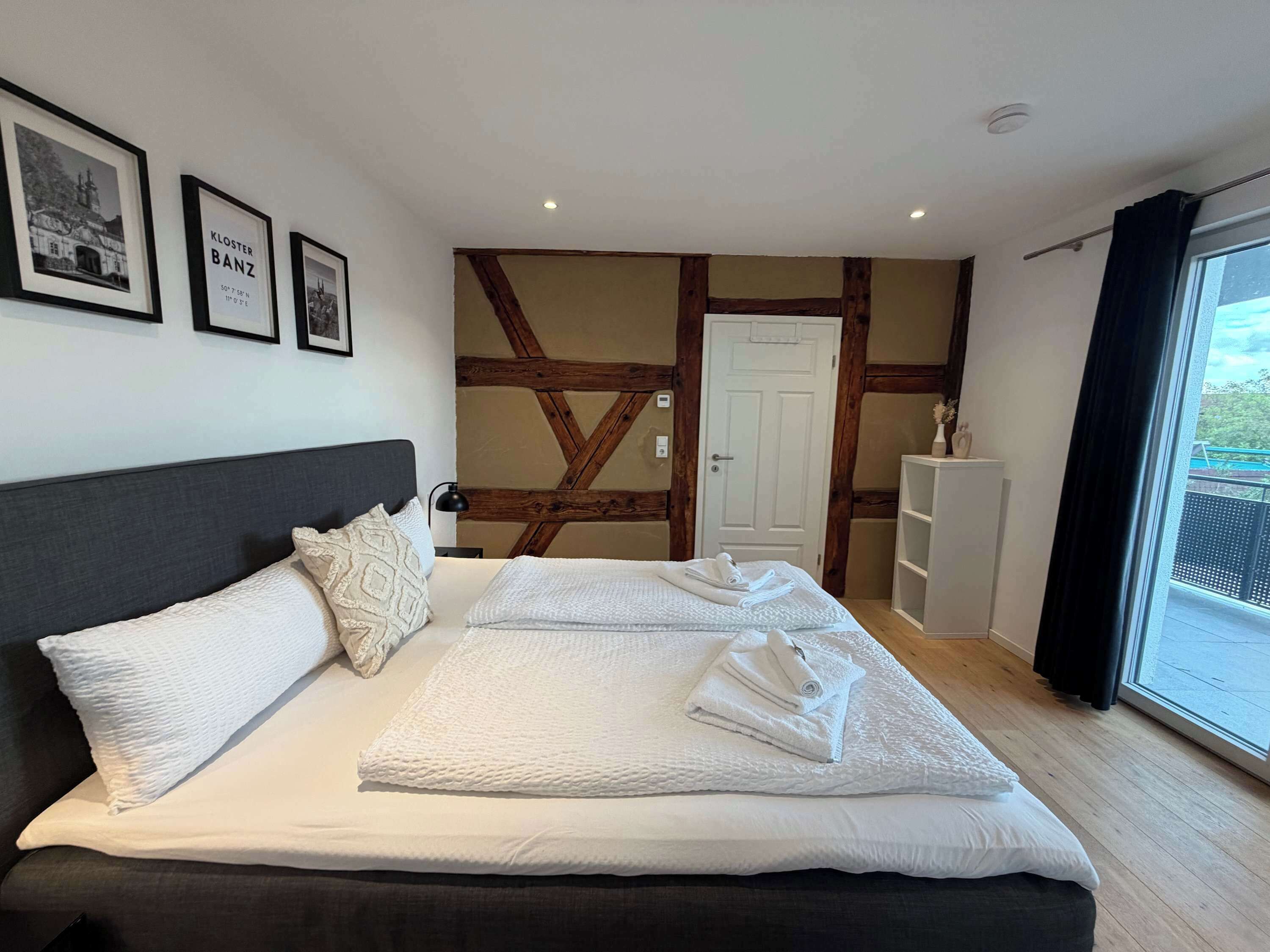A hotel room with a large bed, a headboard, a wooden ceiling, a white door, and a window with a view.