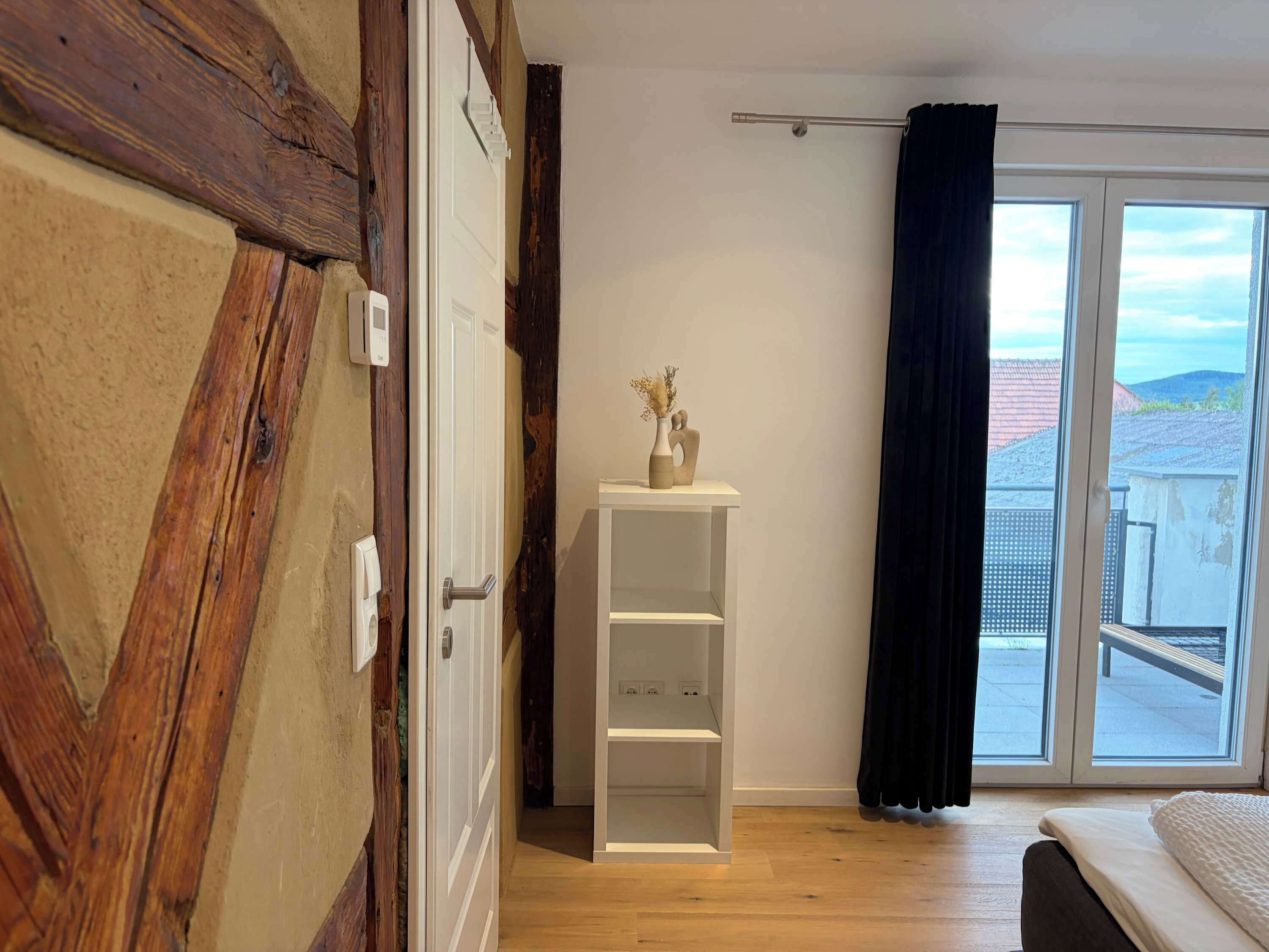 Um quarto com parede de madeira, porta branca, prateleira branca com um vaso, uma cama e vista de um prédio pela janela.