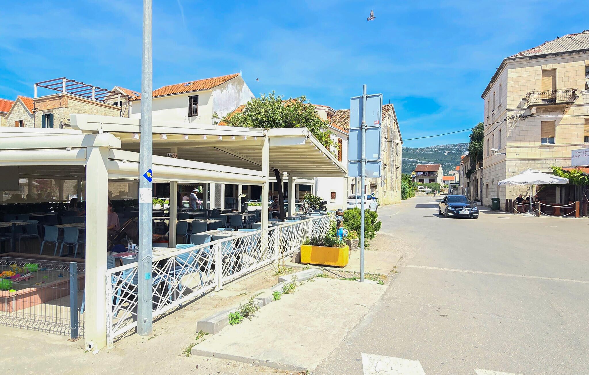 Une vue d’une rue avec une terrasse de restaurant, un bac à fleurs jaune et une voiture garée sur le bord de la route.
