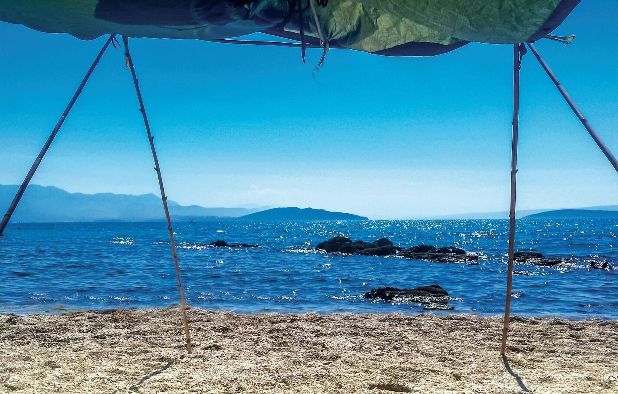 A beach scene with a tent set up on the sand, overlooking a calm sea with visible rocks and distant mountains.