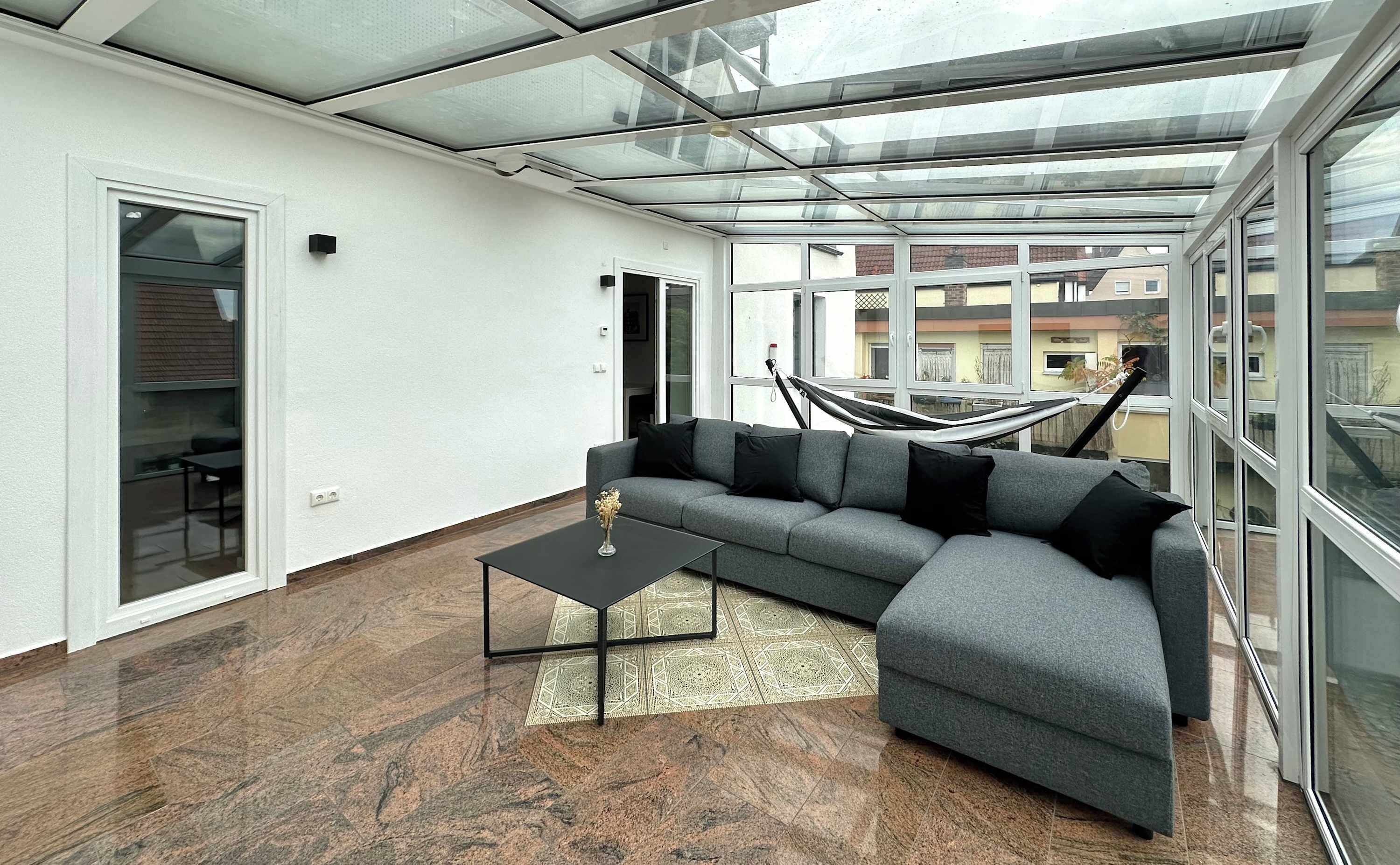 Ein modernes Wohnzimmer mit Glasdecke, einem grauen Ecksofa, einem schwarzen Couchtisch und einem gemusterten Teppich.