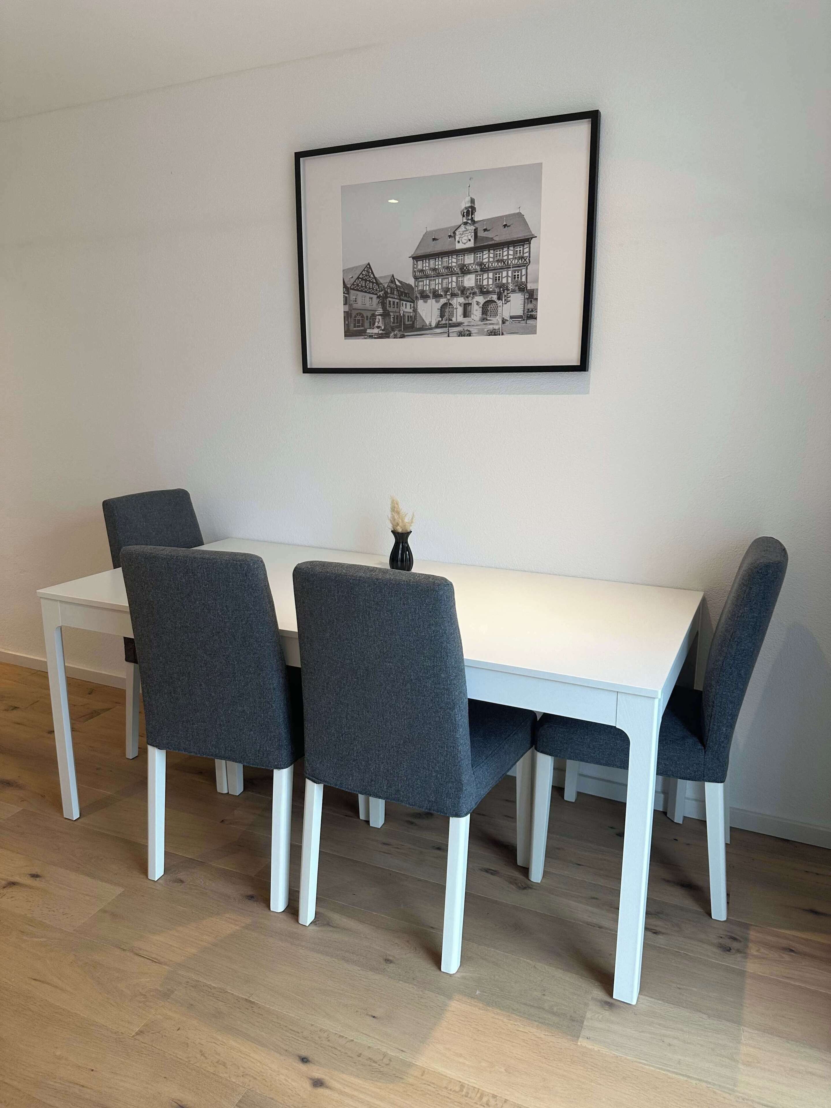 Ein Essbereich mit einem weißen Tisch und vier grauen Stühlen, einem schwarzen und weißen Foto im Rahmen an der Wand und einer Vase mit getrockneten Pflanzen auf dem Tisch.