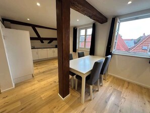 A dining area with a white table and chairs, wooden flooring, and a window with a view of red-roofed buildings.