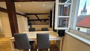 Un coin repas avec une table blanche et quatre chaises grises, une fenêtre donnant sur des immeubles et des poutres en bois apparentes au plafond.