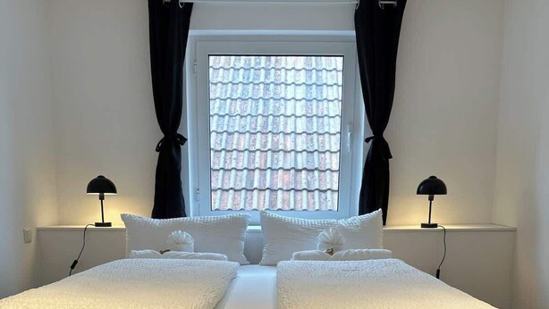 Un lit double avec deux tables de chevet, une fenêtre avec des rideaux et une vue sur un toit recouvert de tuiles.