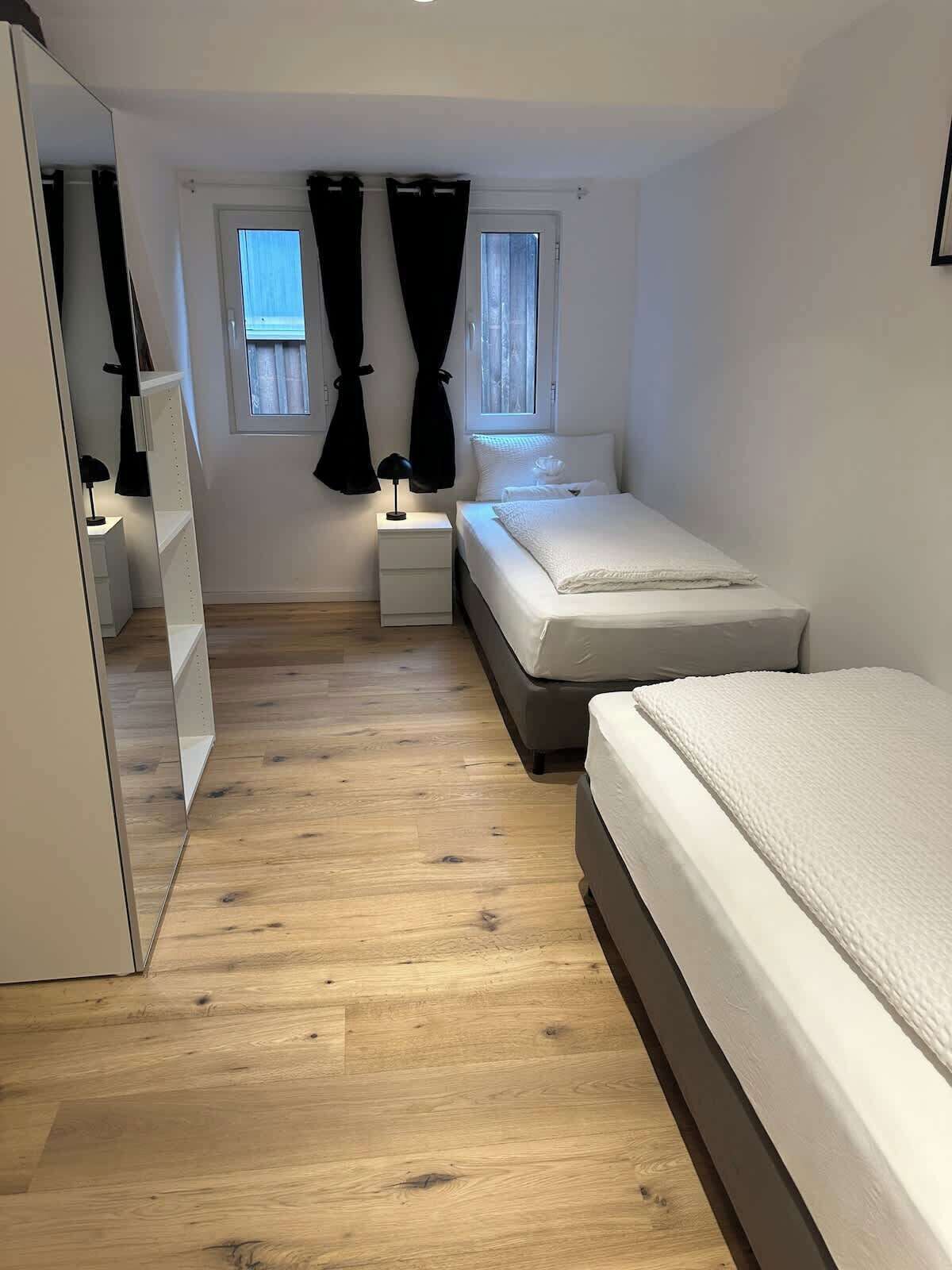 Une chambre avec un plancher en bois, deux lits, une table de chevet, un miroir et deux fenêtres avec des rideaux.