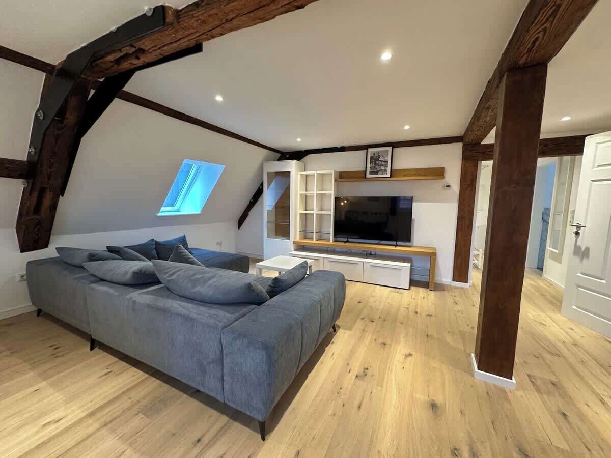 A loft-style room with exposed wooden beams, a grey sofa, a TV, and a window.