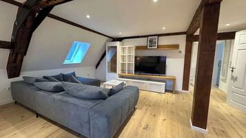 A loft-style room with exposed wooden beams, a grey sofa, a TV, and a window.