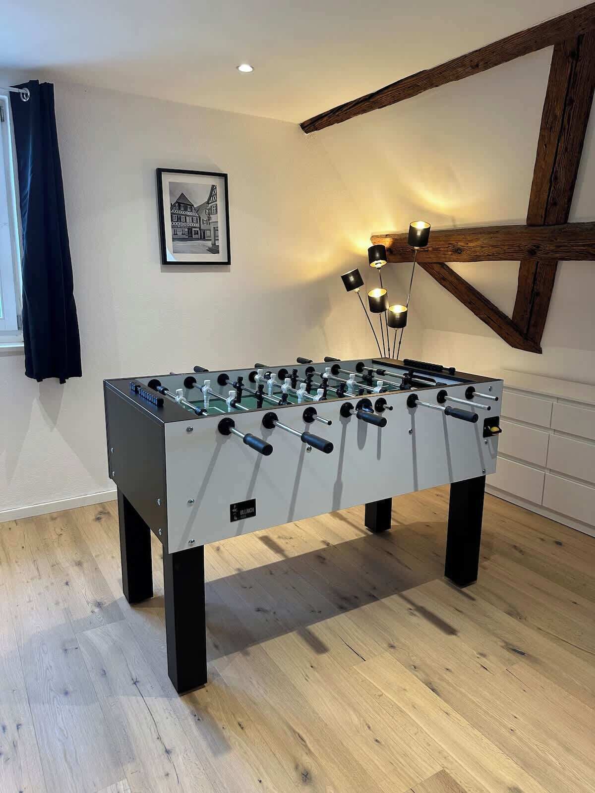 A foosball table in a room with wooden flooring, a framed picture on the wall, and a window with a curtain.