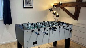 Une table de baby-foot dans une pièce dont le sol est recouvert de parquet, un tableau encadré au mur et une fenêtre avec un rideau.