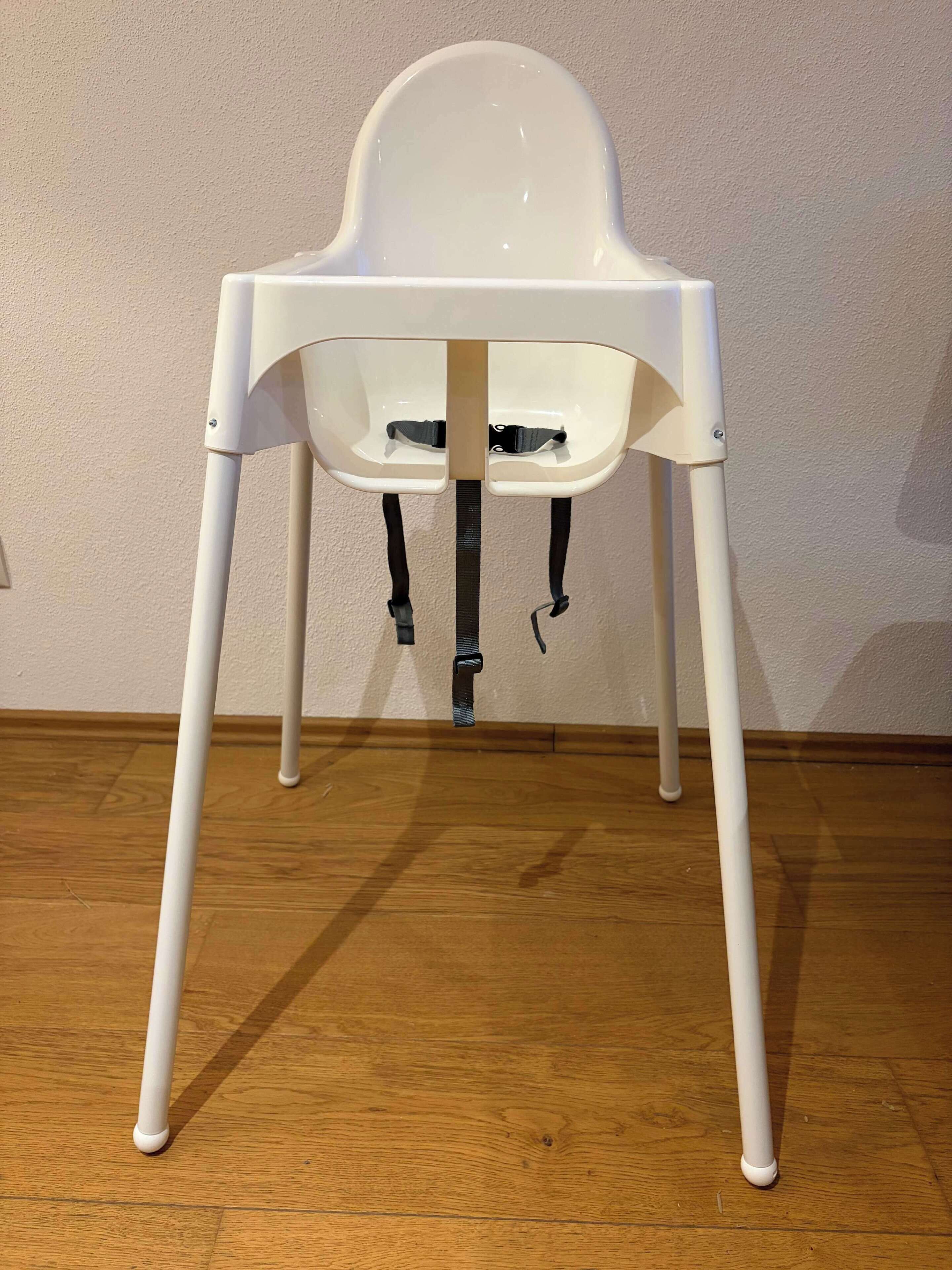 A white high chair with a tray, positioned against a wall with a wooden floor.