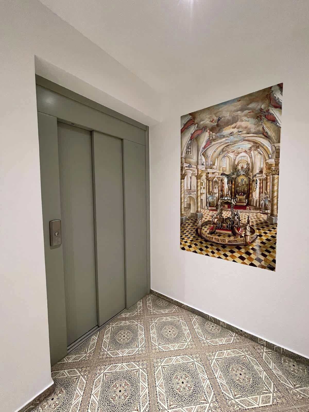 Ein Flur mit einem gemusterten Boden, einer Tür und einem an der Wand hängenden Kunstwerk, das einen prunkvollen Innenraum zeigt.