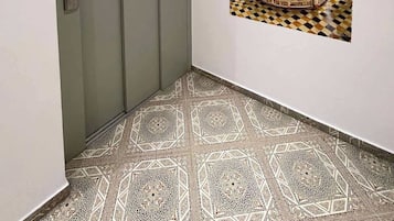 Een gang met een patroon in de vloer, een deur en een wanddecoratie waarop een versierd interieur te zien is.