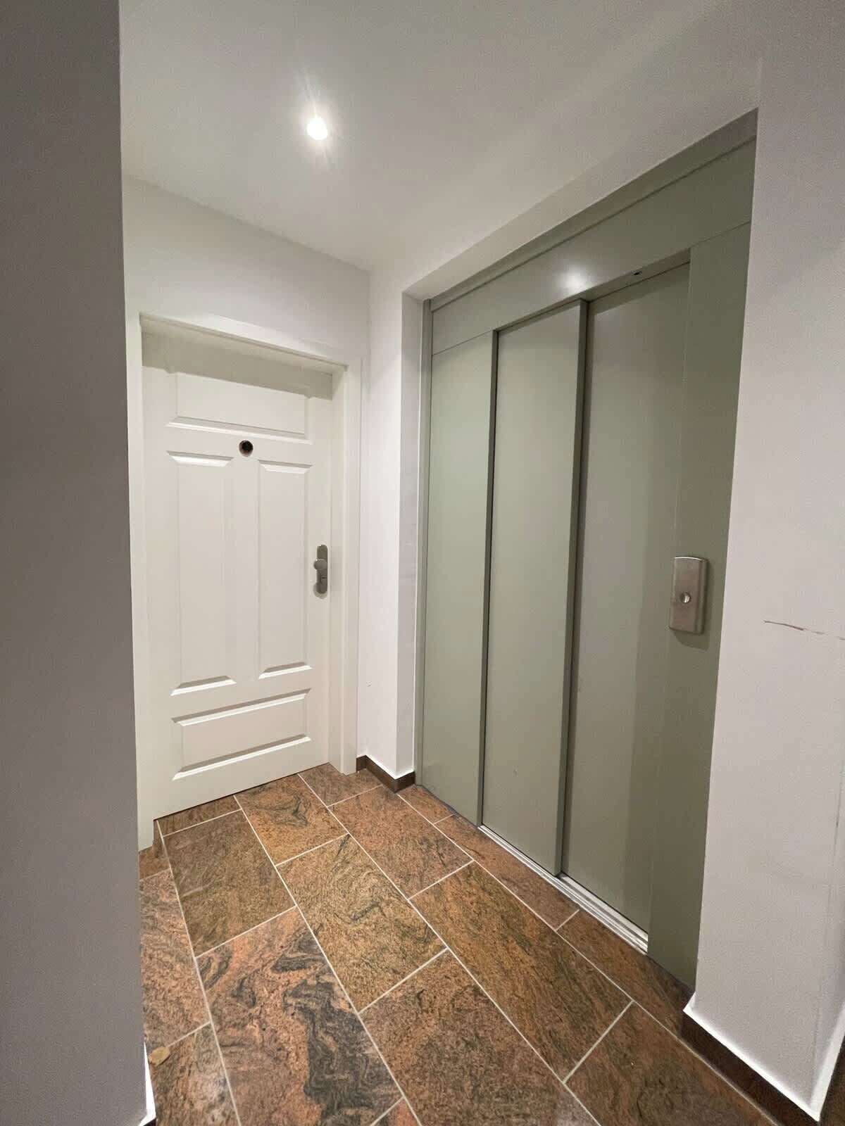 A hallway with a white door and a sliding glass door leading to a room. The floor is covered with brown patterned tiles.