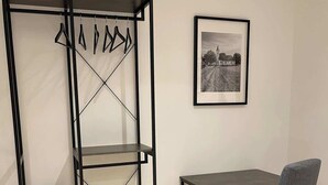 Una habitación con una estantería metálica, una pequeña mesa, una silla y un cuadro en la pared.