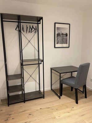 Ein Zimmer mit einem Metallregal, einem kleinen Tisch, einem Stuhl und einem gerahmten Bild an der Wand.