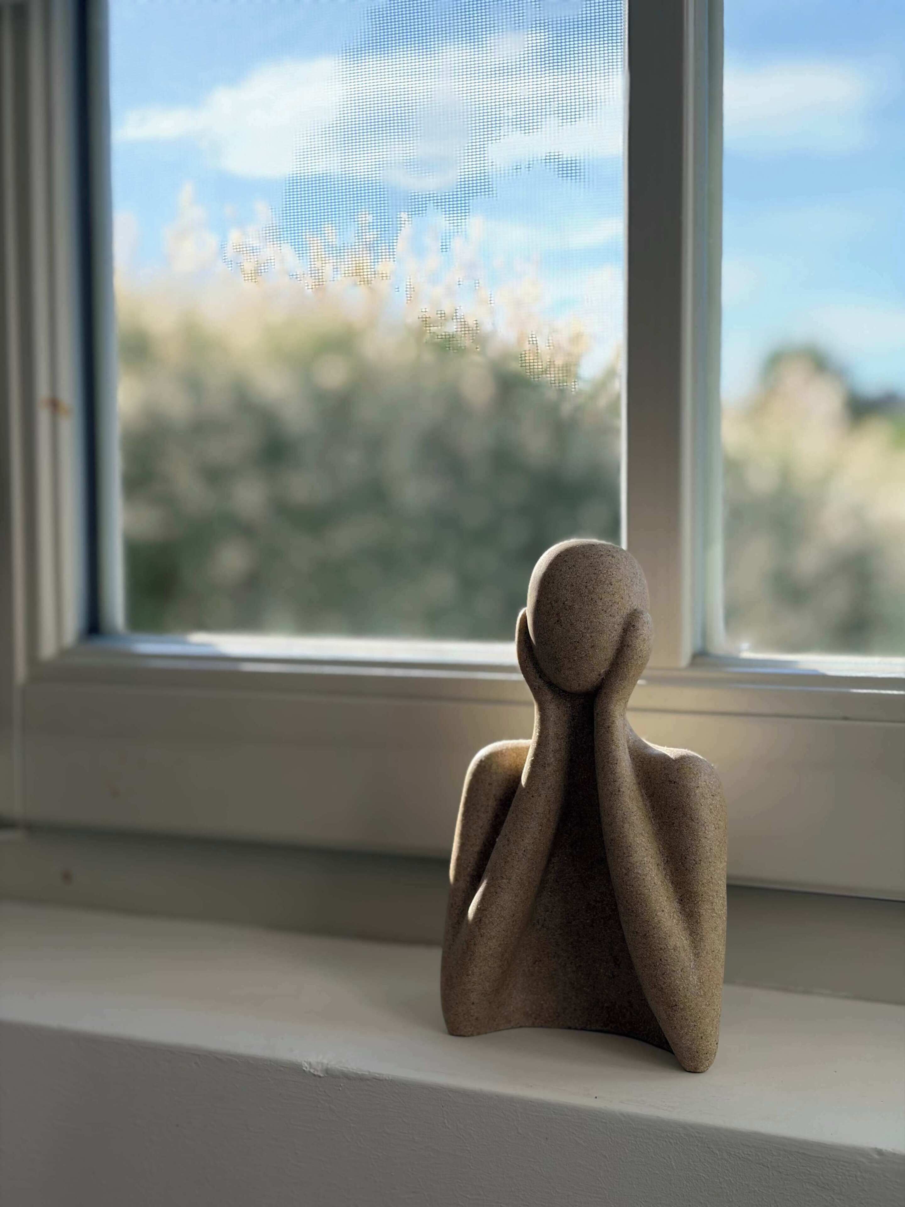 Una escultura en un alféizar con vistas a un cielo nublado y follaje exterior.