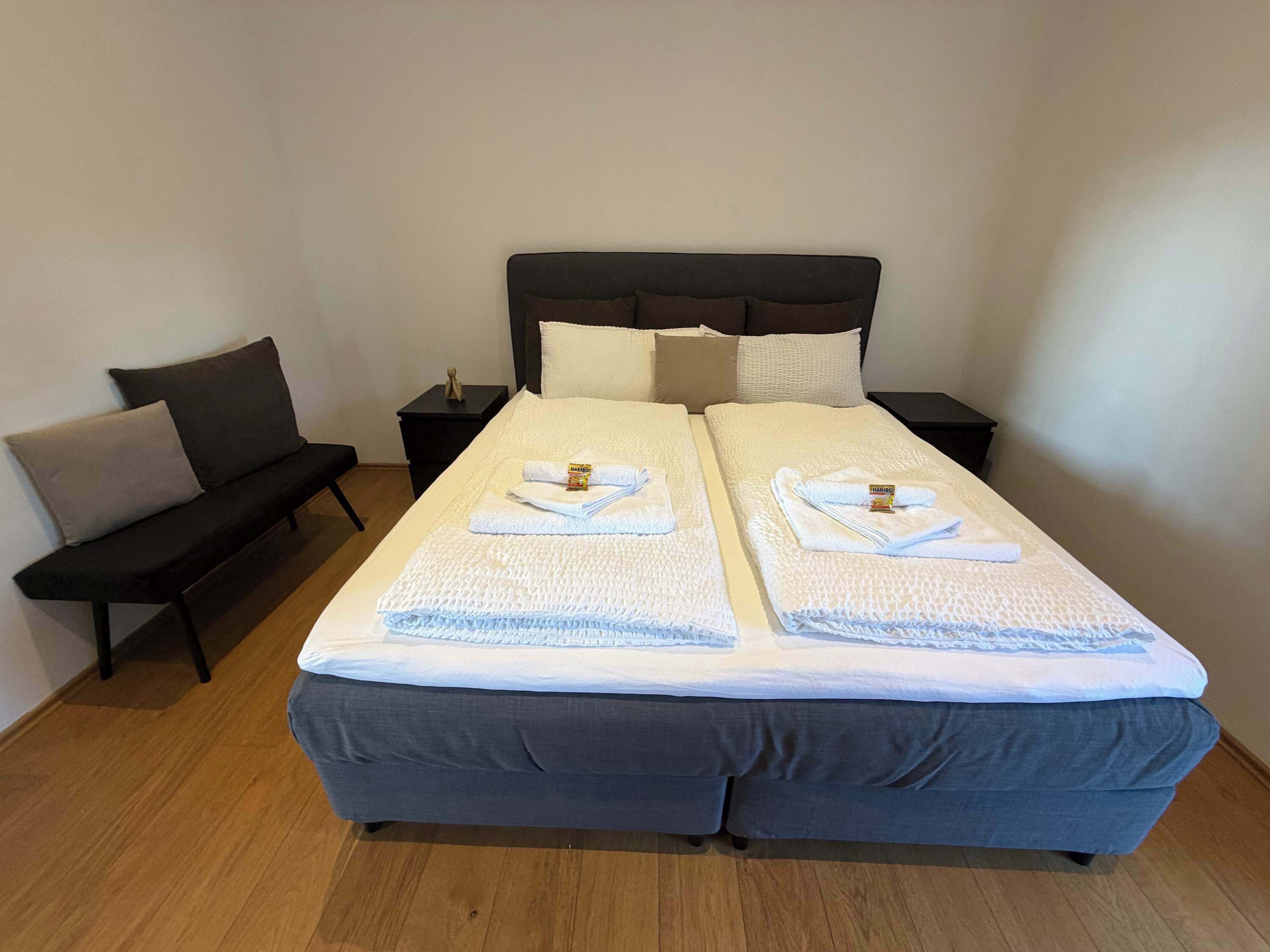 Ein Doppelbett mit zwei Kissen, ein Nachttisch, ein kleiner Beistelltisch und eine Bank mit Sitzkissen.
